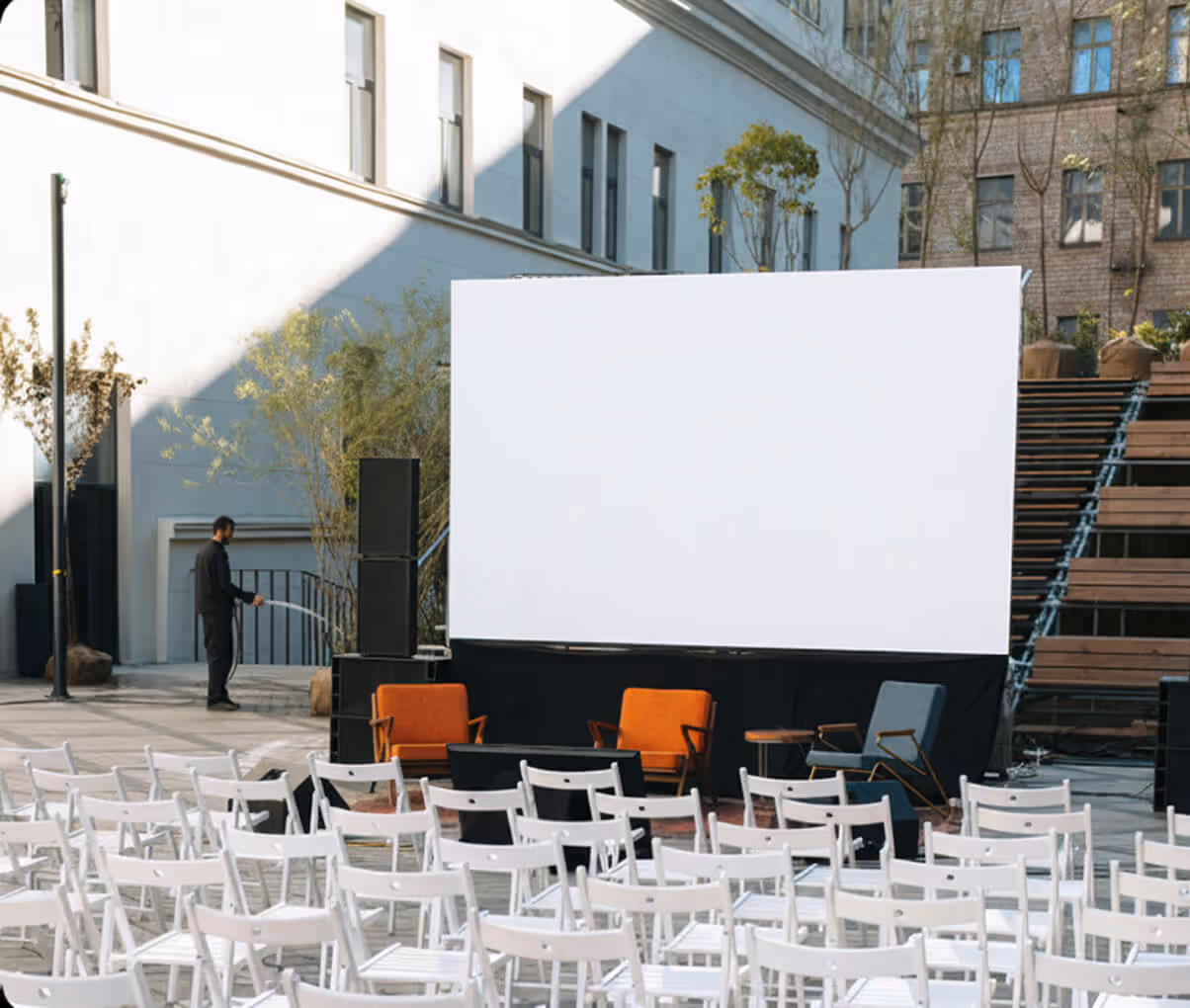Outdoor event setup with white folding chairs, empty large screen, and three armchairs on stage.