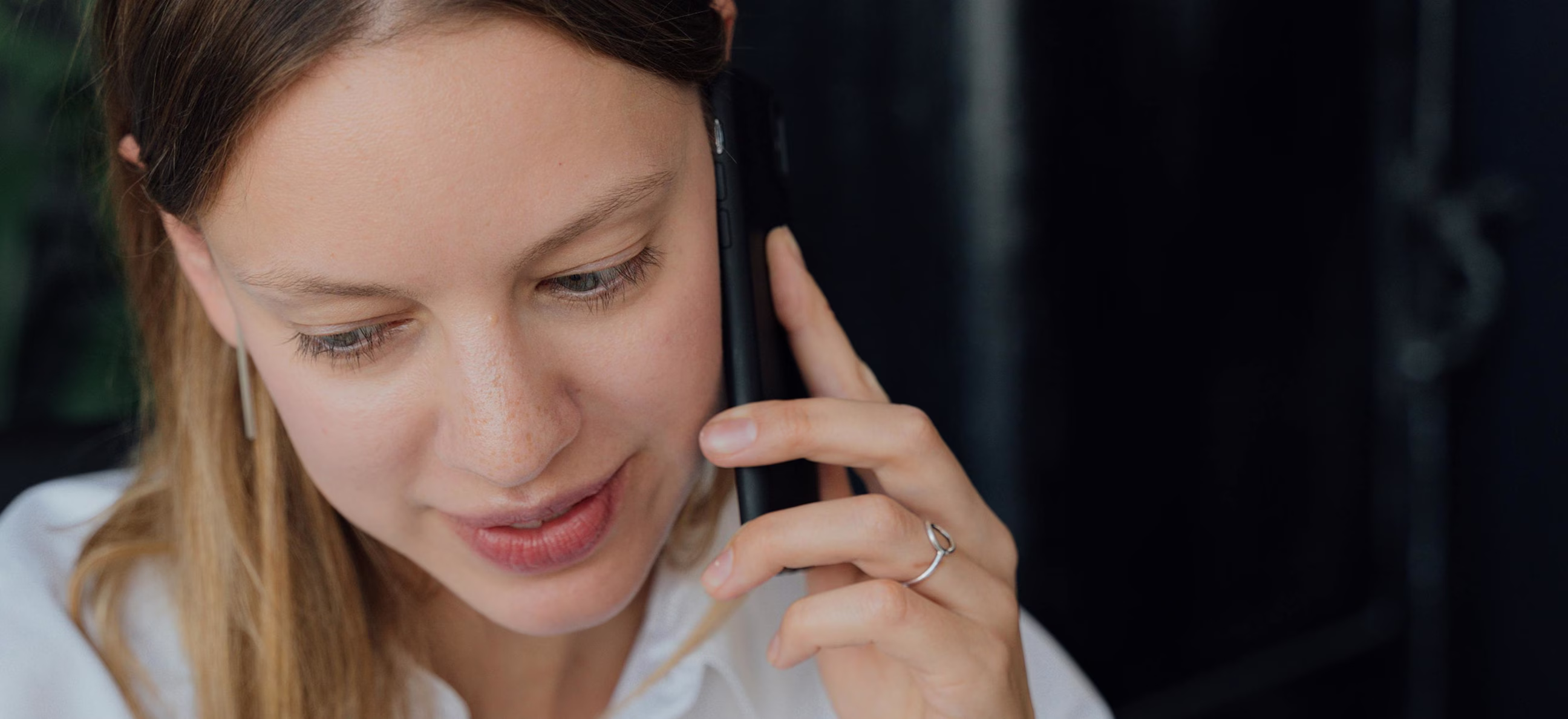 Close-up of a woman speaking on a black phone, wearing a white top and a silver ring.