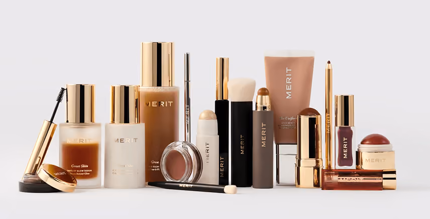 Array of Merit brand makeup products including foundation, lipstick, blush, and brushes on white background.