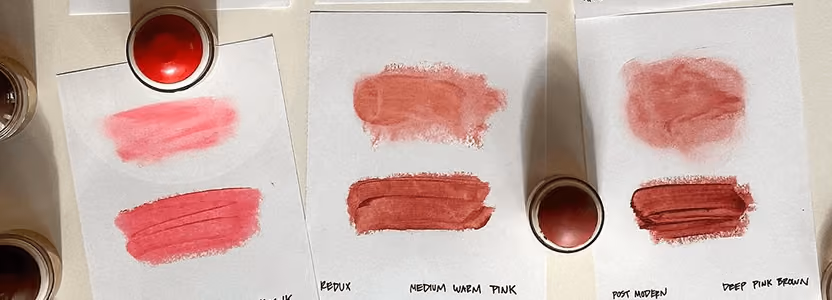 Samples of four shades of pink lipstick with smudges on white paper and containers above them.