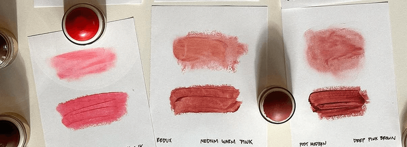Samples of four shades of pink lipstick with smudges on white paper and containers above them.