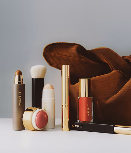 Collection of Merit brand makeup products including lipstick, blush, brush, and nail polish with brown cloth.