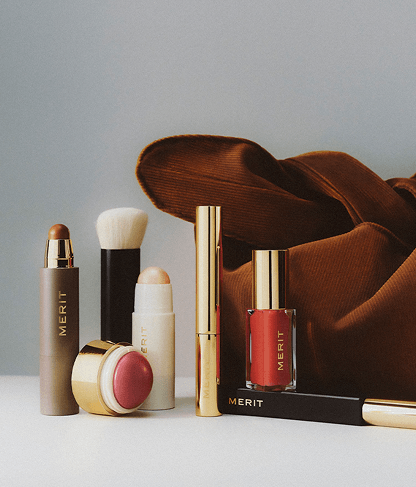 Collection of Merit brand makeup products including lipstick, blush, brush, and nail polish with brown cloth.