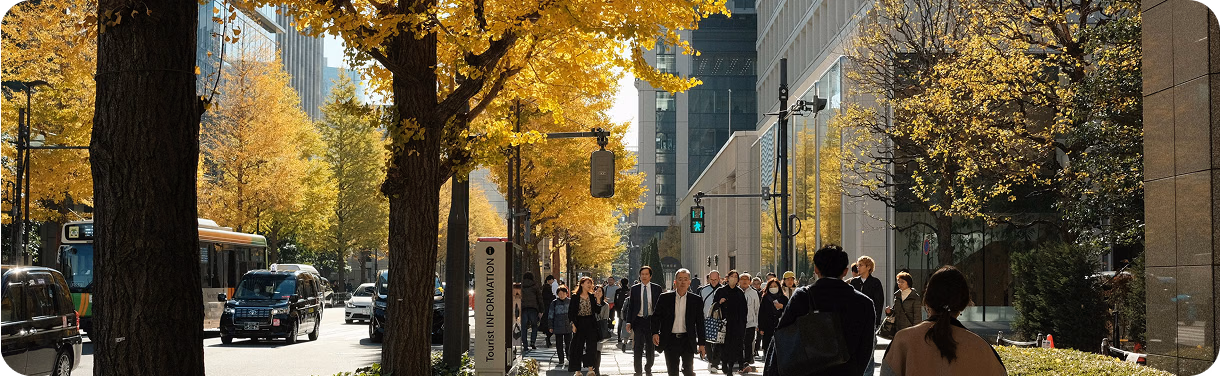 Busy city sidewalk lined with golden autumn trees and pedestrians in business attire.
