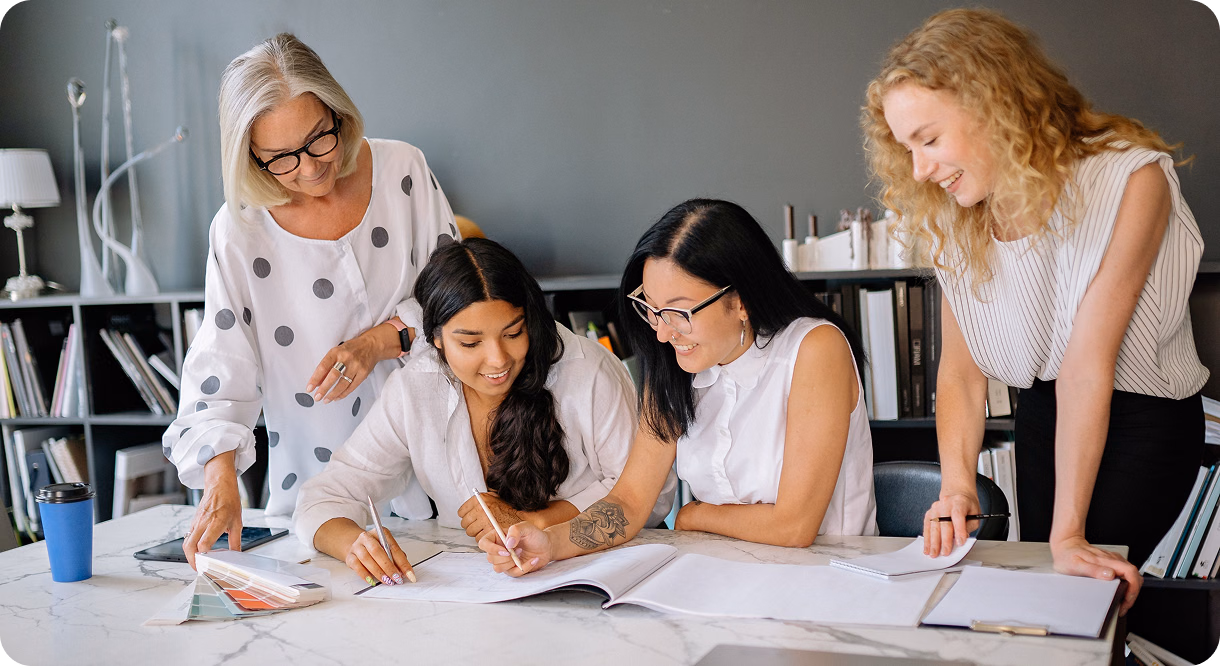 Four women gathered around a table reviewing papers and planning together in an office.