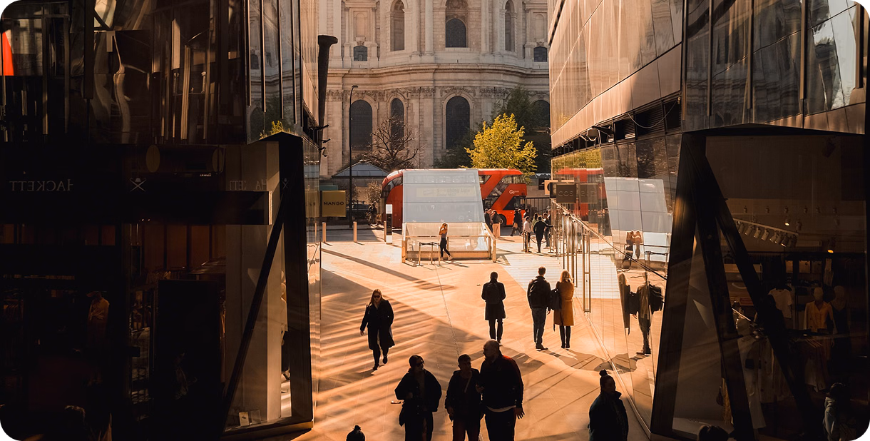 Commuters walking through a sunlit city plaza between office buildings toward a domed cathedral.
