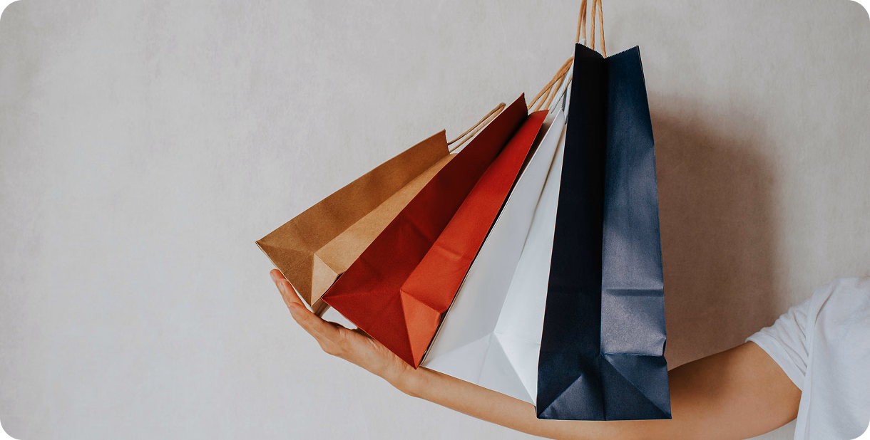 Hand holding several colorful paper shopping bags against a light wall.