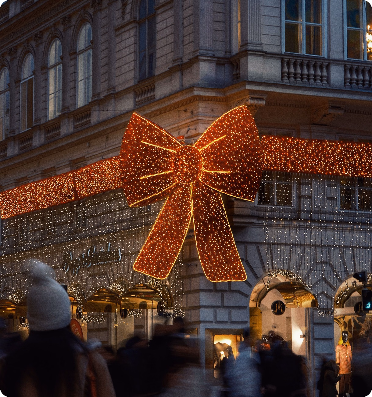 Large illuminated red holiday bow and string lights decorating a storefront at dusk.