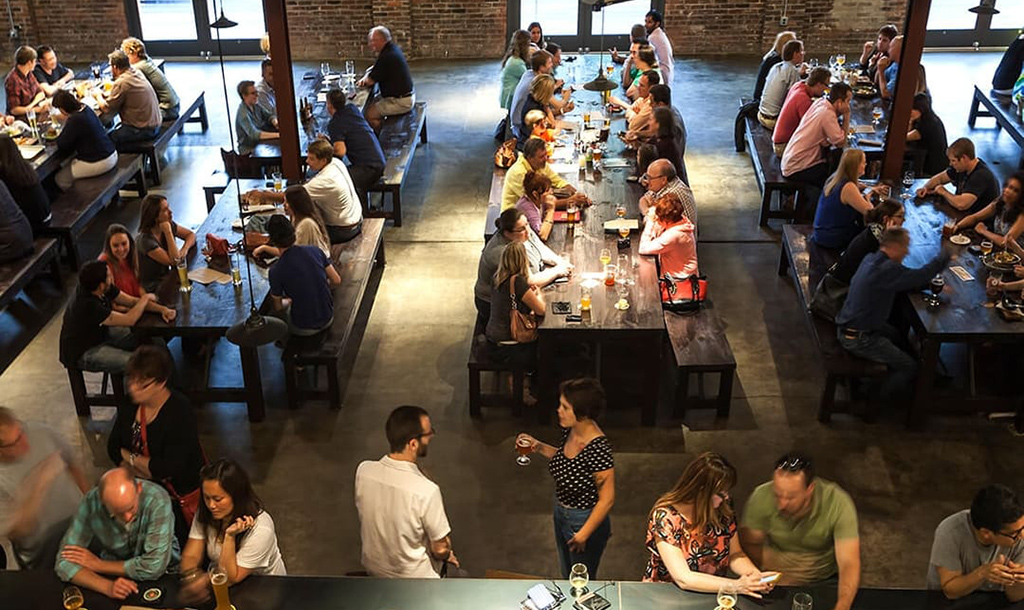 Large open Bierhall with bar and long table filled will people enjoying themselves