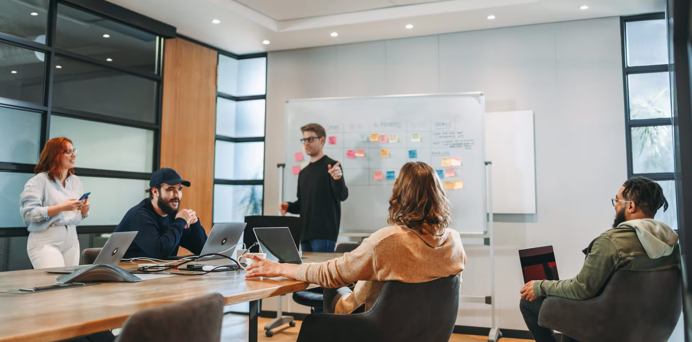 Team meeting in a modern office, a man at a whiteboard of sticky notes leading a discussion with colleagues at laptops
