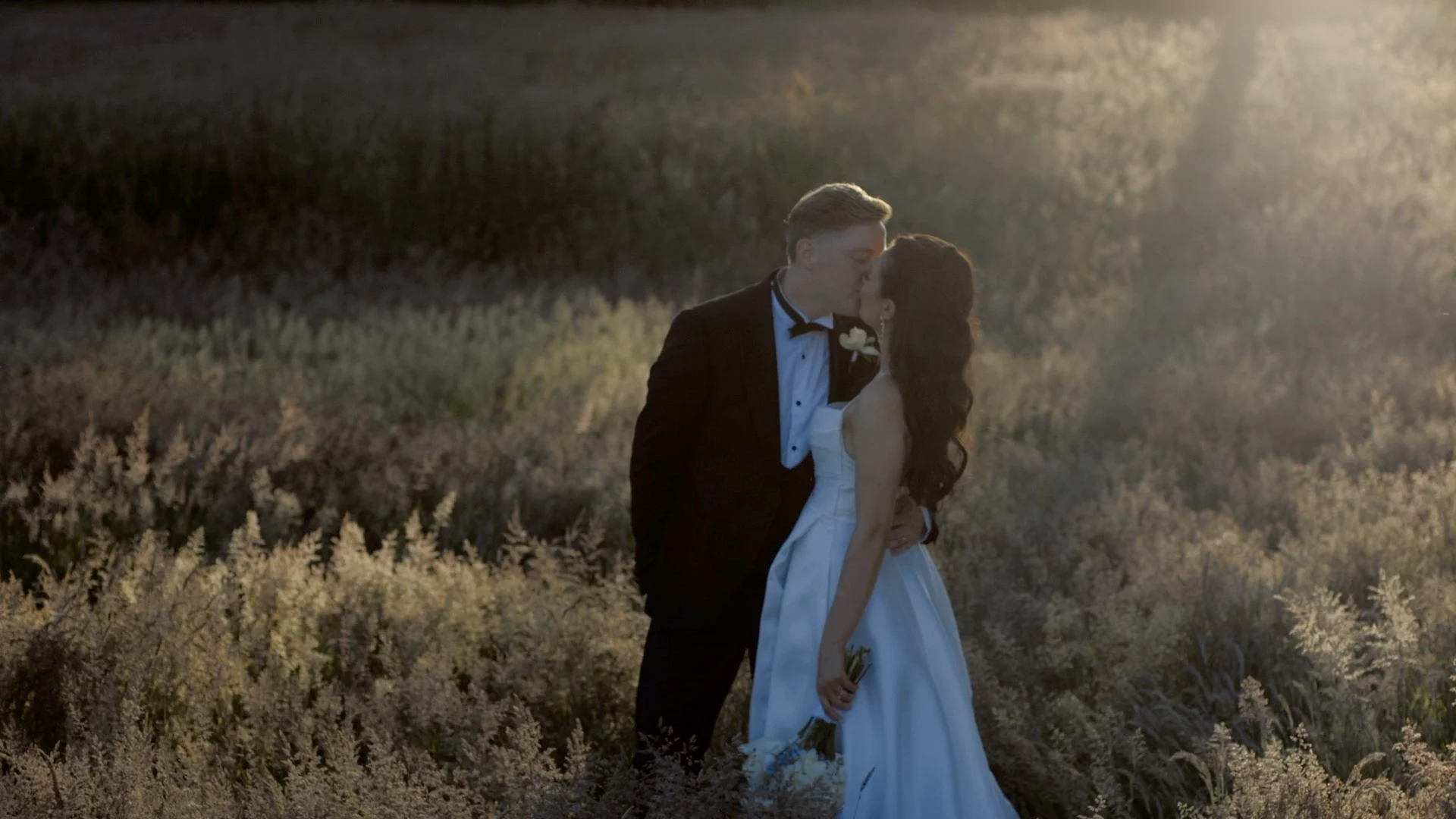 Close-up of bride and groom sitting on a vehicle, holding hands with the bride in a beaded white wedding dress and the groom in a brown jacket.
