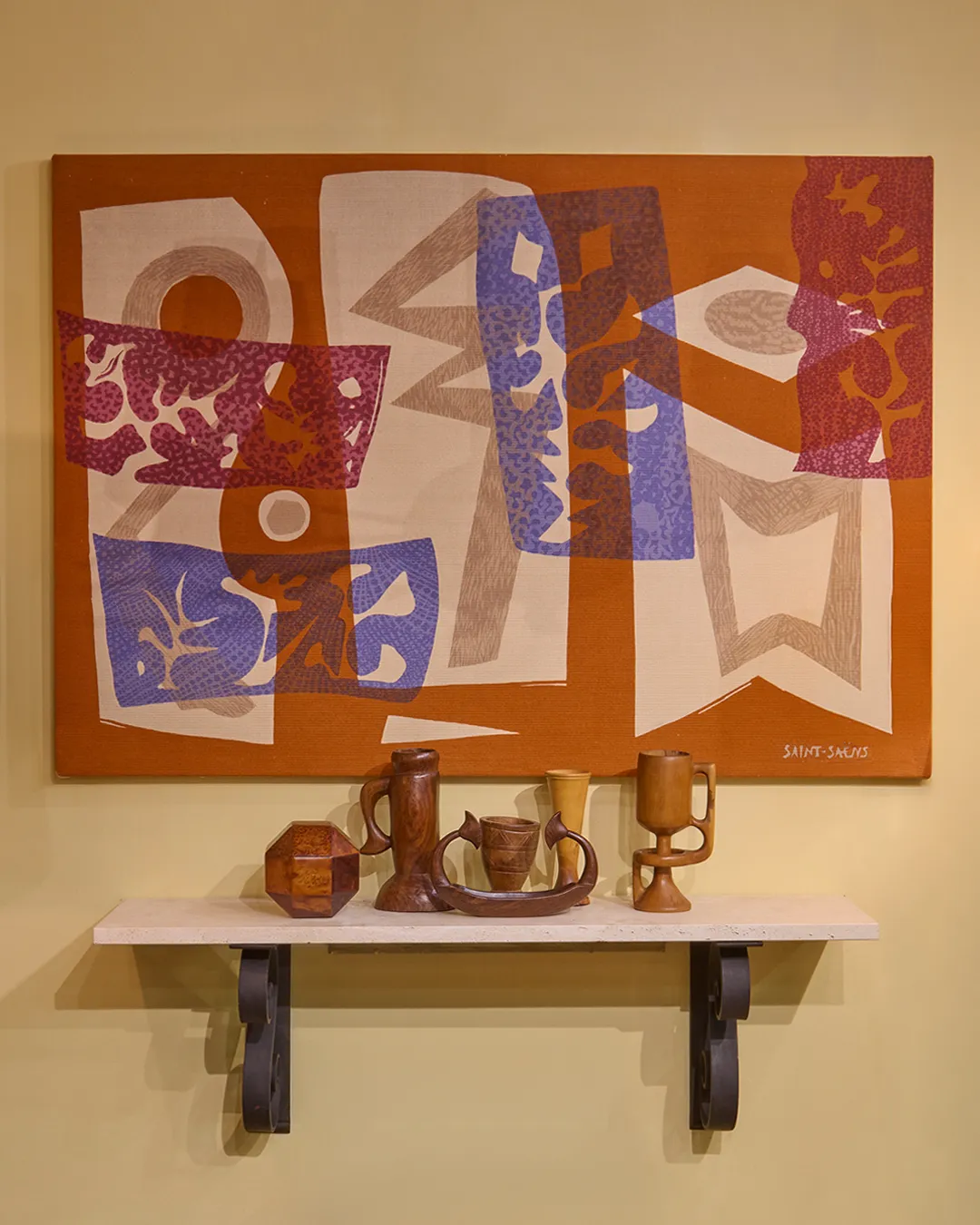Abstract wall art with geometric shapes in rust, purple, white, and blue above a shelf holding various wooden carved vessels and containers.