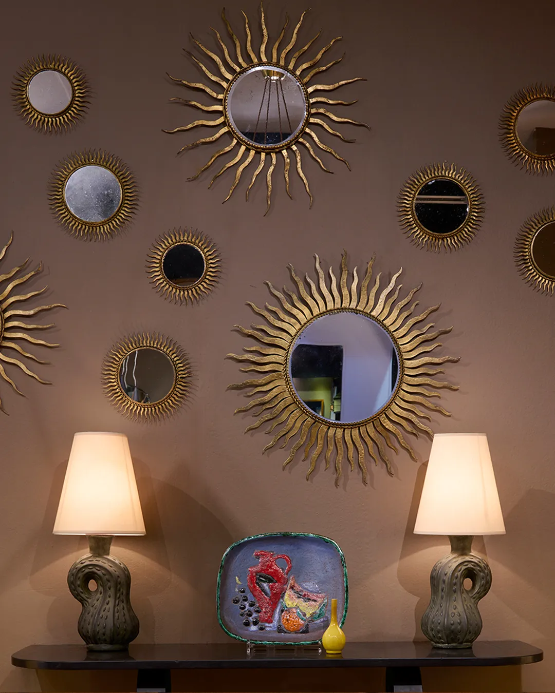 Wall decorated with multiple round sunburst mirrors above a console table holding two sculptural lamps and a colorful decorative plate with a small yellow vase.