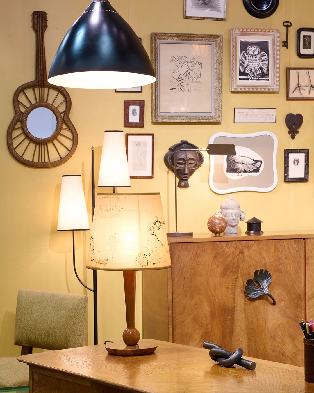 Cozy room corner with wooden desk, table lamps, floor lamp, a green chair, wall art, and decorative sculptures on a wooden cabinet.