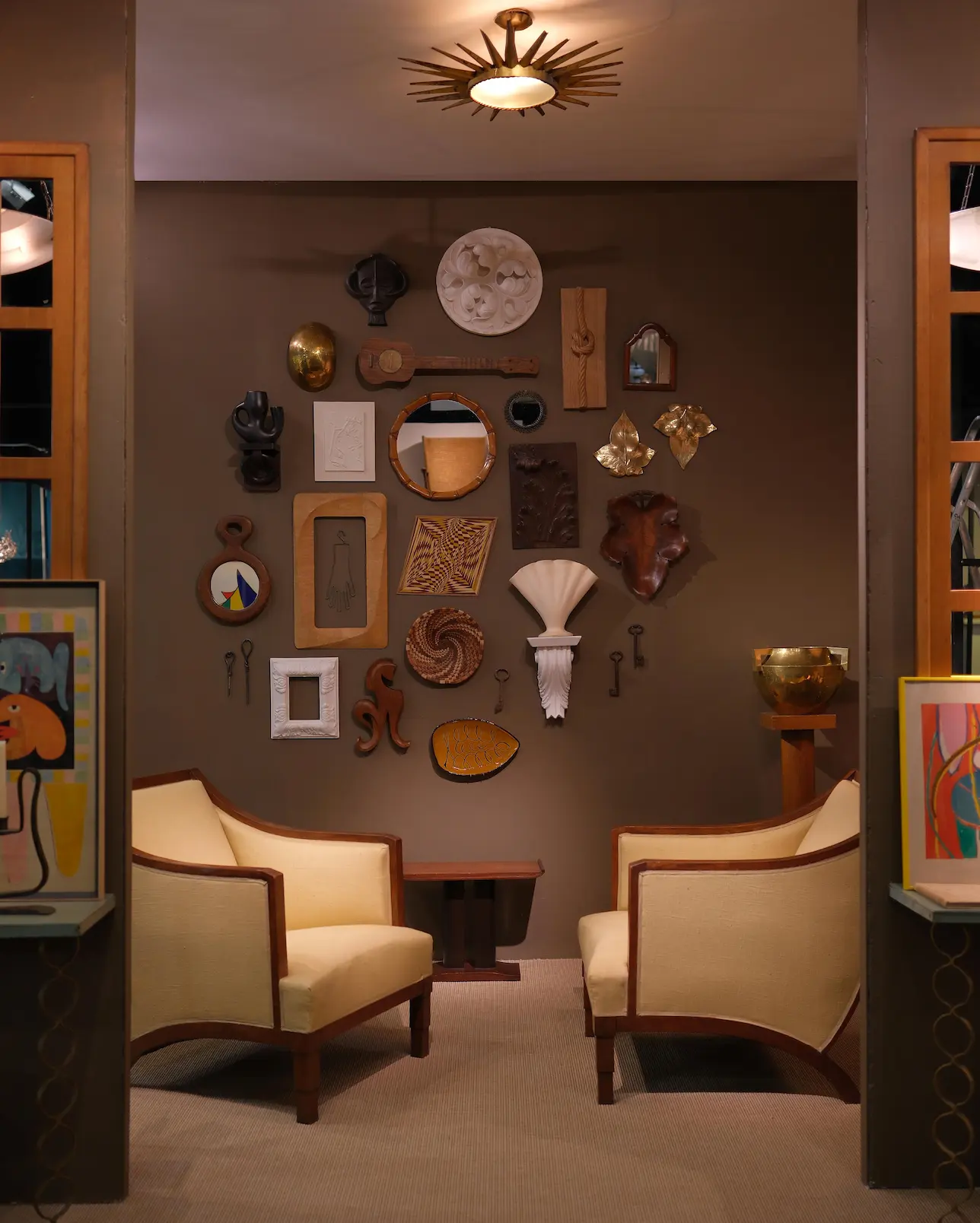 Cozy seating area with two beige armchairs facing a wall decorated with eclectic wooden and metal art pieces and mirrors.