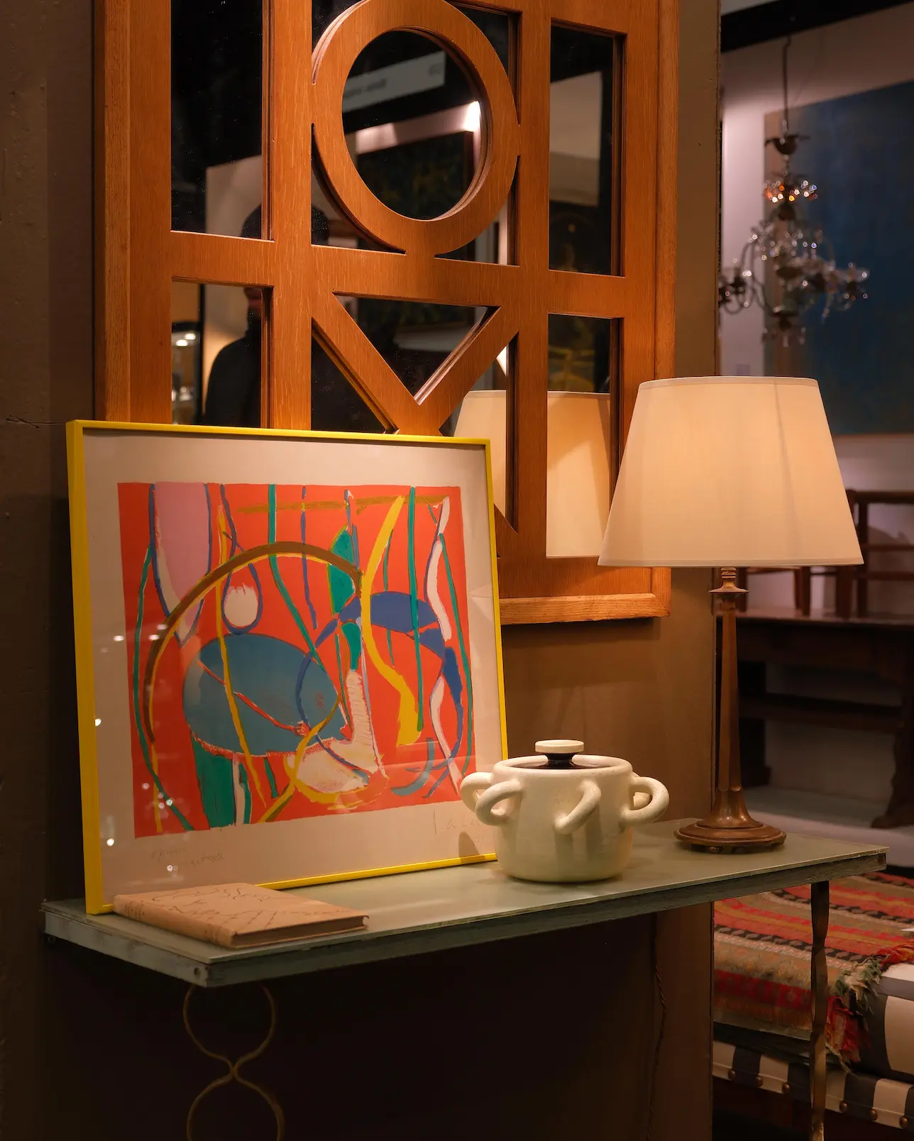 Decorative shelf with a colorful abstract framed artwork, a ceramic pot, and a lit table lamp against a wooden framed mirror.