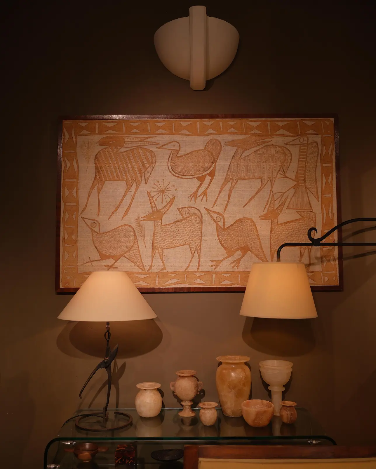 Wall hanging with abstract animal and bird designs above a glass table holding stone pottery and two beige lamps.