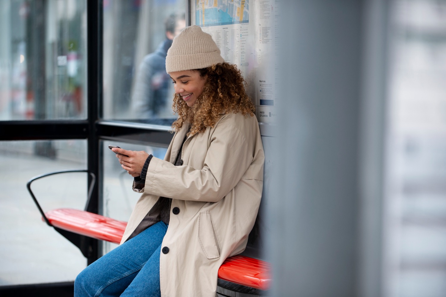Lady. in woolly hat and coat sitting in bus stop smiling at phone 