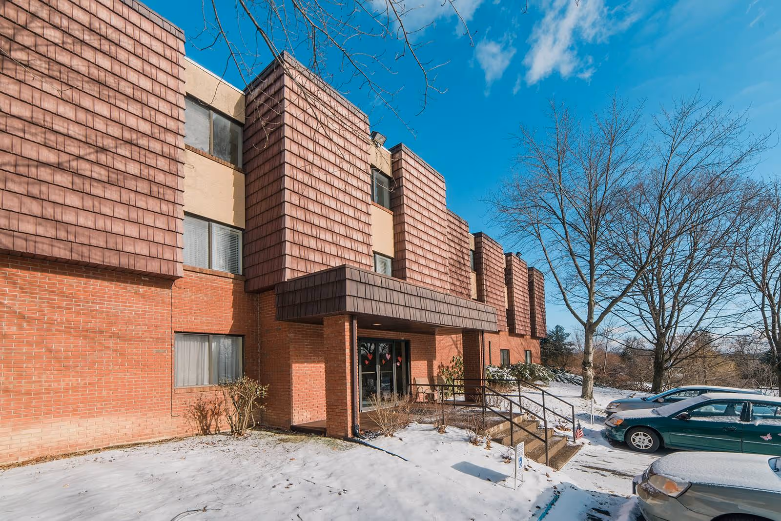 Snow-covered ground and parked cars outside a multi-story brick and shingle building with leafless trees under a blue sky.