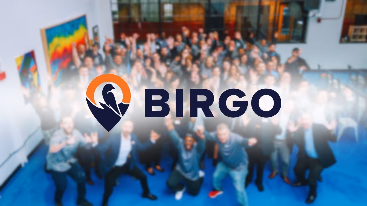 Blurred group of people standing indoors with a large BIRGO logo featuring a stylized bird and orange circle overlayed in the center.