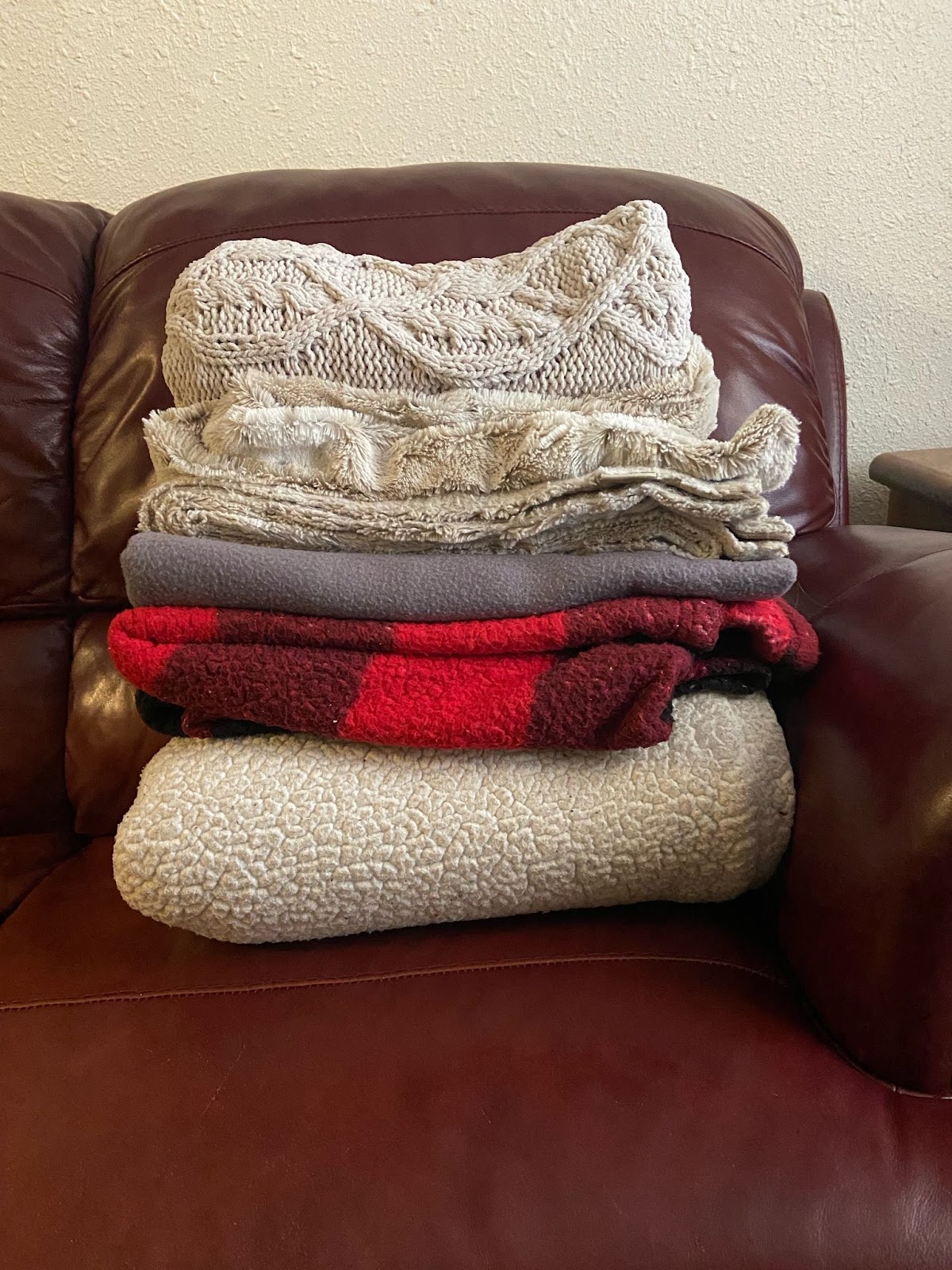 one way to make your apartment cozy for the fall is to get cozy blankets. This image shows 5 blankets folded n top of one another and they are sitting on top of a brown leather couch cushion.