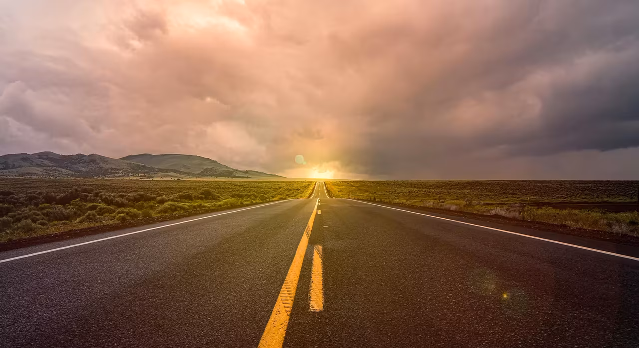 An empty road stretches into the distance, with a vibrant sunset casting warm colors in the sky behind it. 