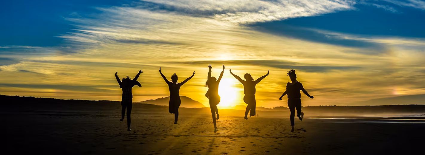 A cheerful group of people jumps in unison on a beach, surrounded by sand and the ocean under a bright sky.