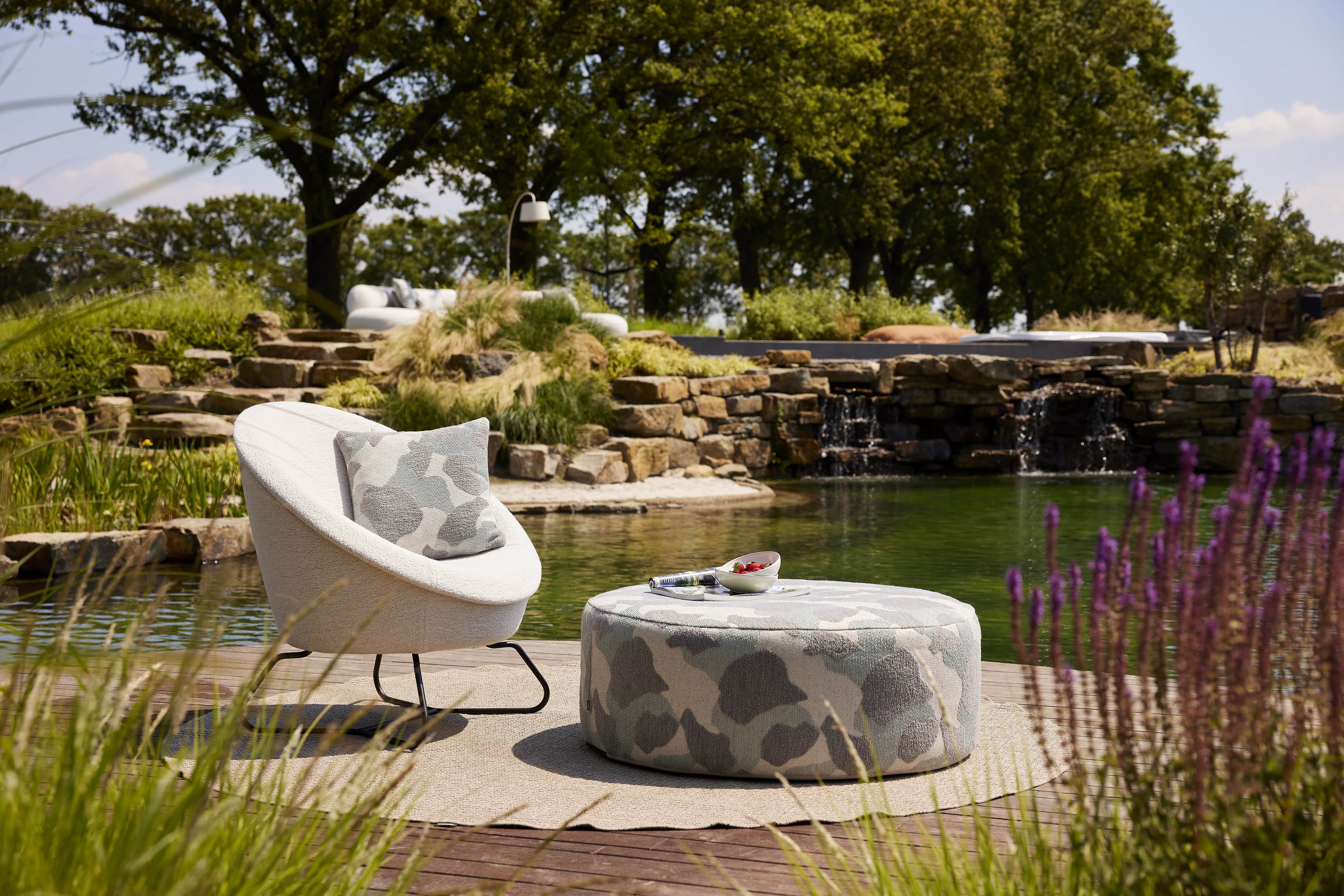 Moderne stoel en poef bij een groene vijver met waterval en lavendel