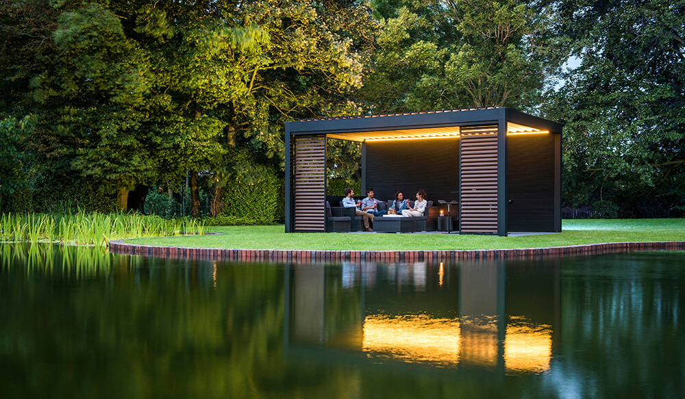 Renson lamellenoverkapping bij vijver met verlichte zitruimte en groene omgeving