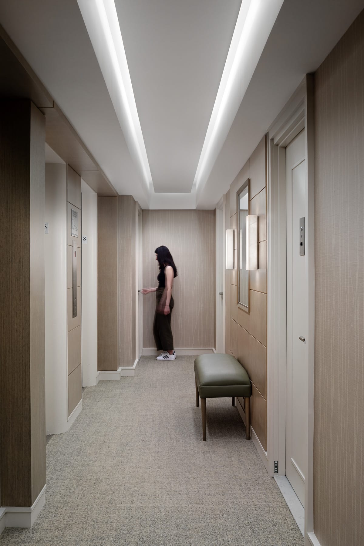 Person in minimalist elevator lobby with beige walls, wall sconces, and a green bench.
