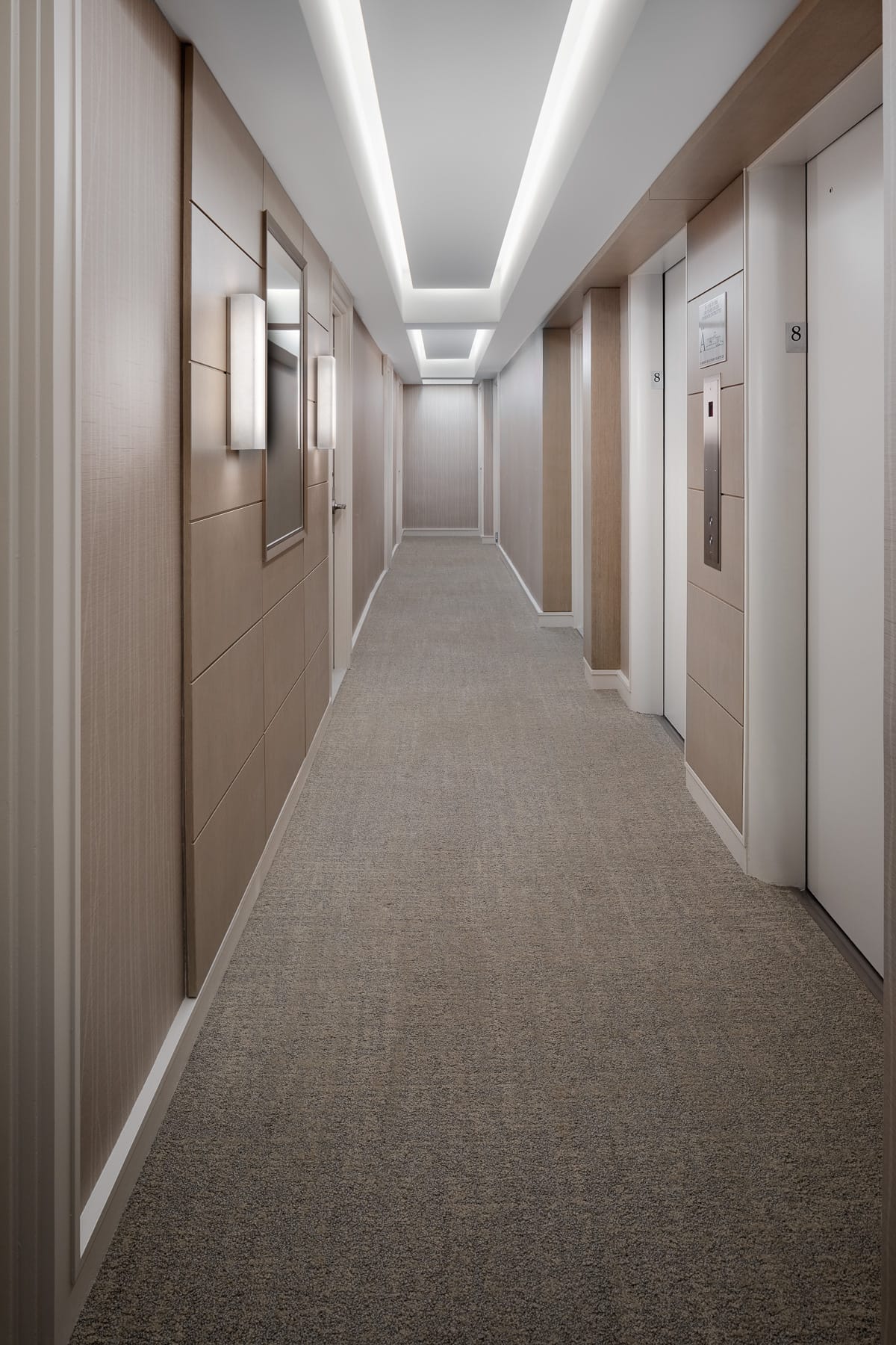 Neutral-toned elevator hallway with wood panels, carpet, and recessed ceiling lighting.