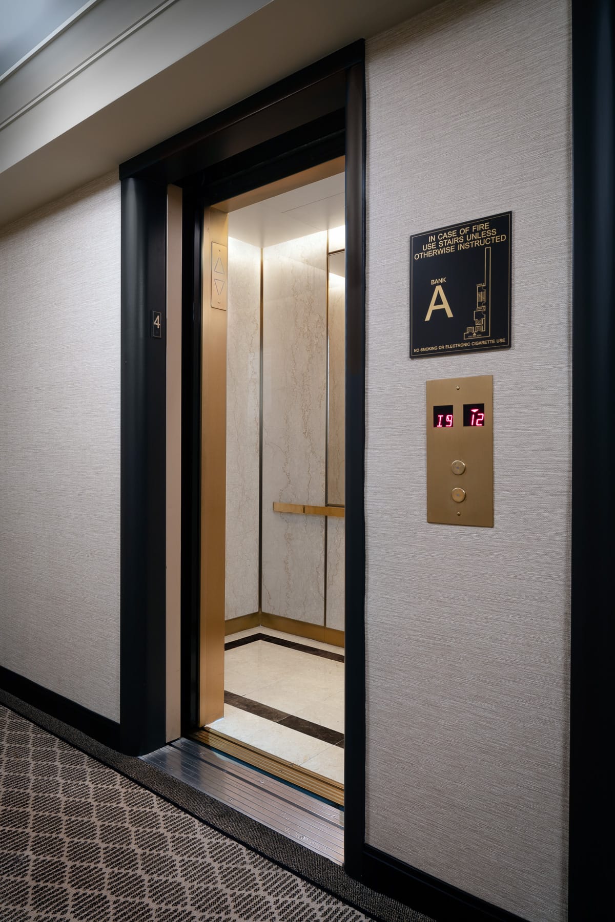 An open elevator on the fourth floor reveals a light stone interior beside call buttons and a fire-safety sign marked “Bank A.”