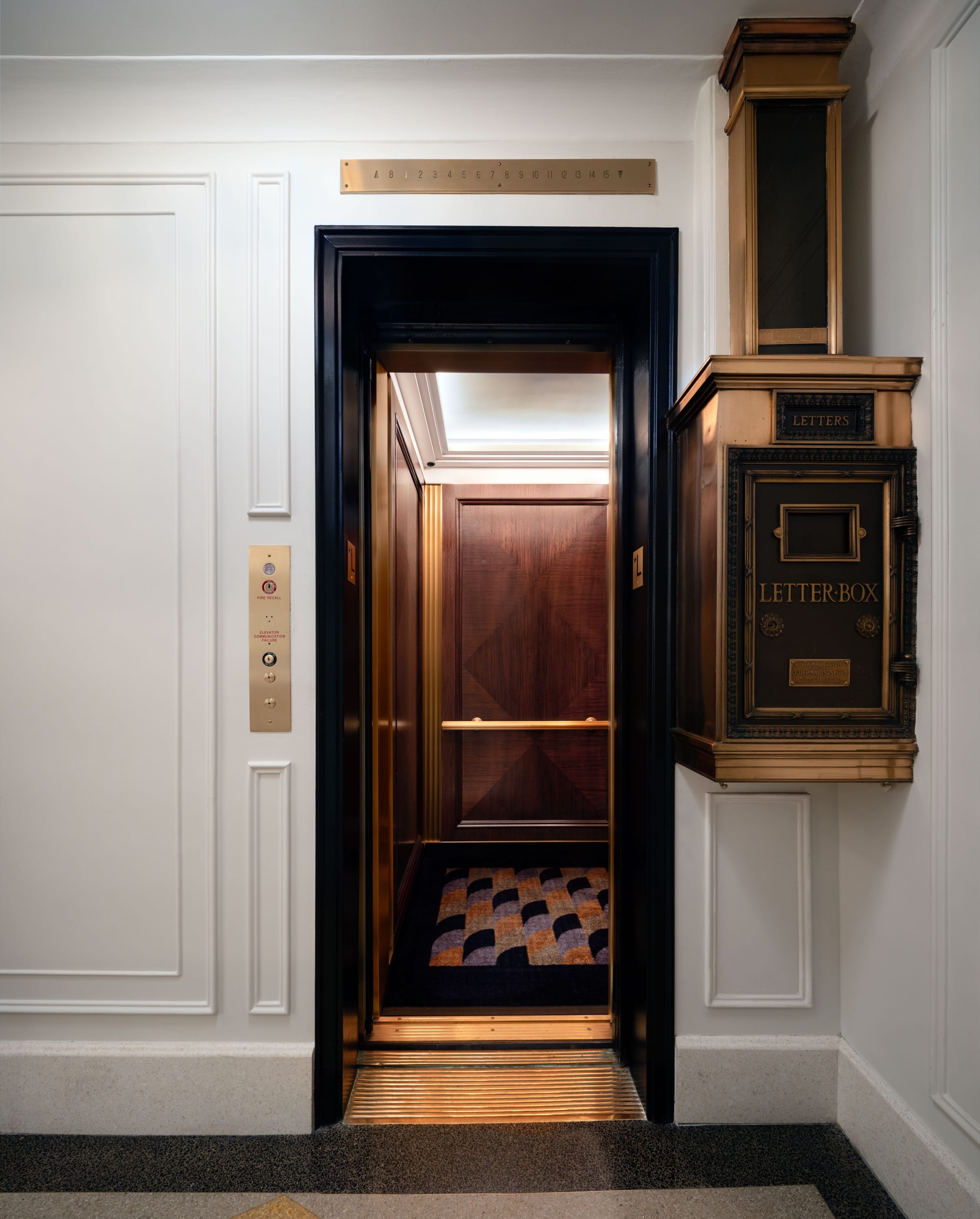 An open elevator with dark wood paneling, brass trim, patterned carpet, and an adjacent ornate wall-mounted letter box in a white-paneled hallway.