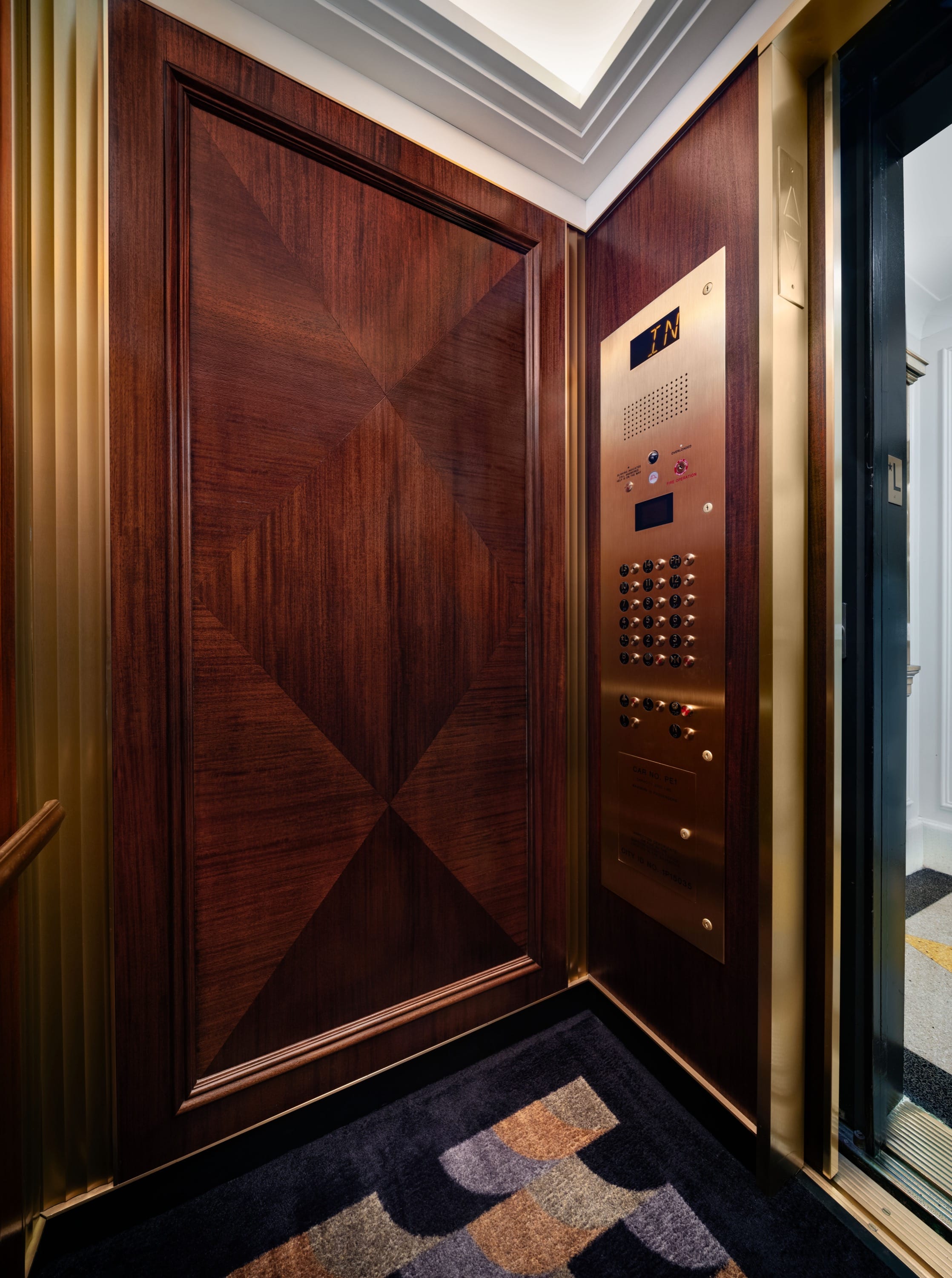 Inside the elevator, geometric dark wood panels surround a brass control panel with floor buttons and a patterned carpet under a recessed ceiling light.