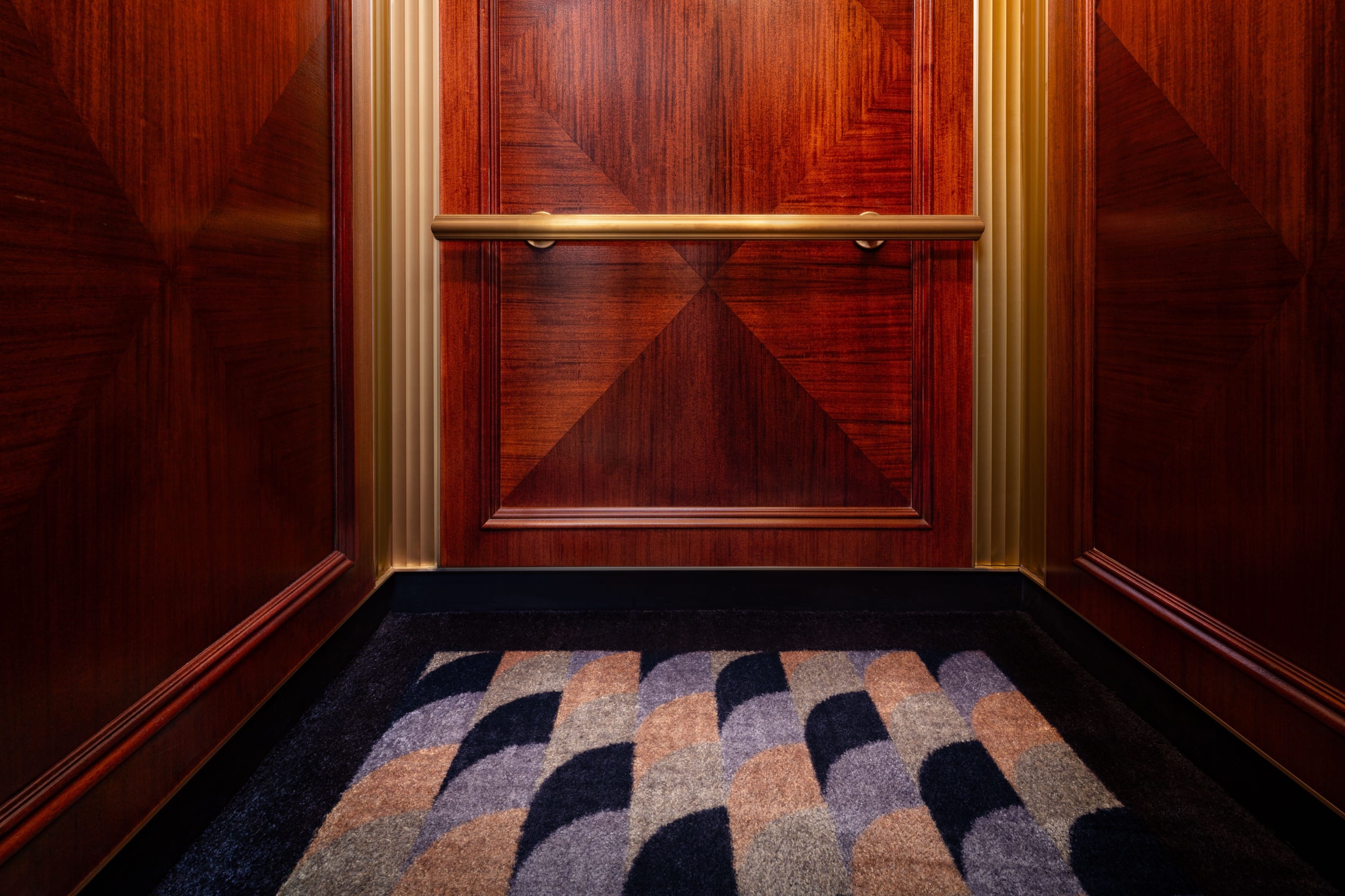Lower interior of the elevator with geometric wood paneling, a horizontal brass handrail, and a multicolored geometric-patterned carpet.