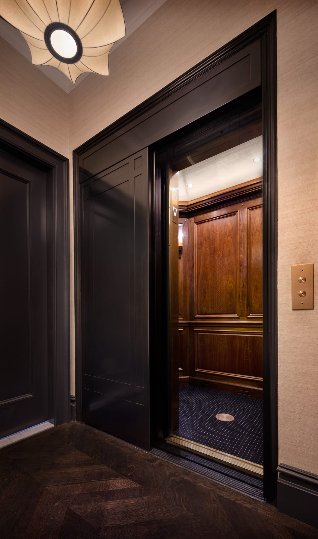 An open dark-paneled elevator reveals a wood-paneled interior with a patterned carpet, set in a hallway with dark trim and a wall-mounted call button.