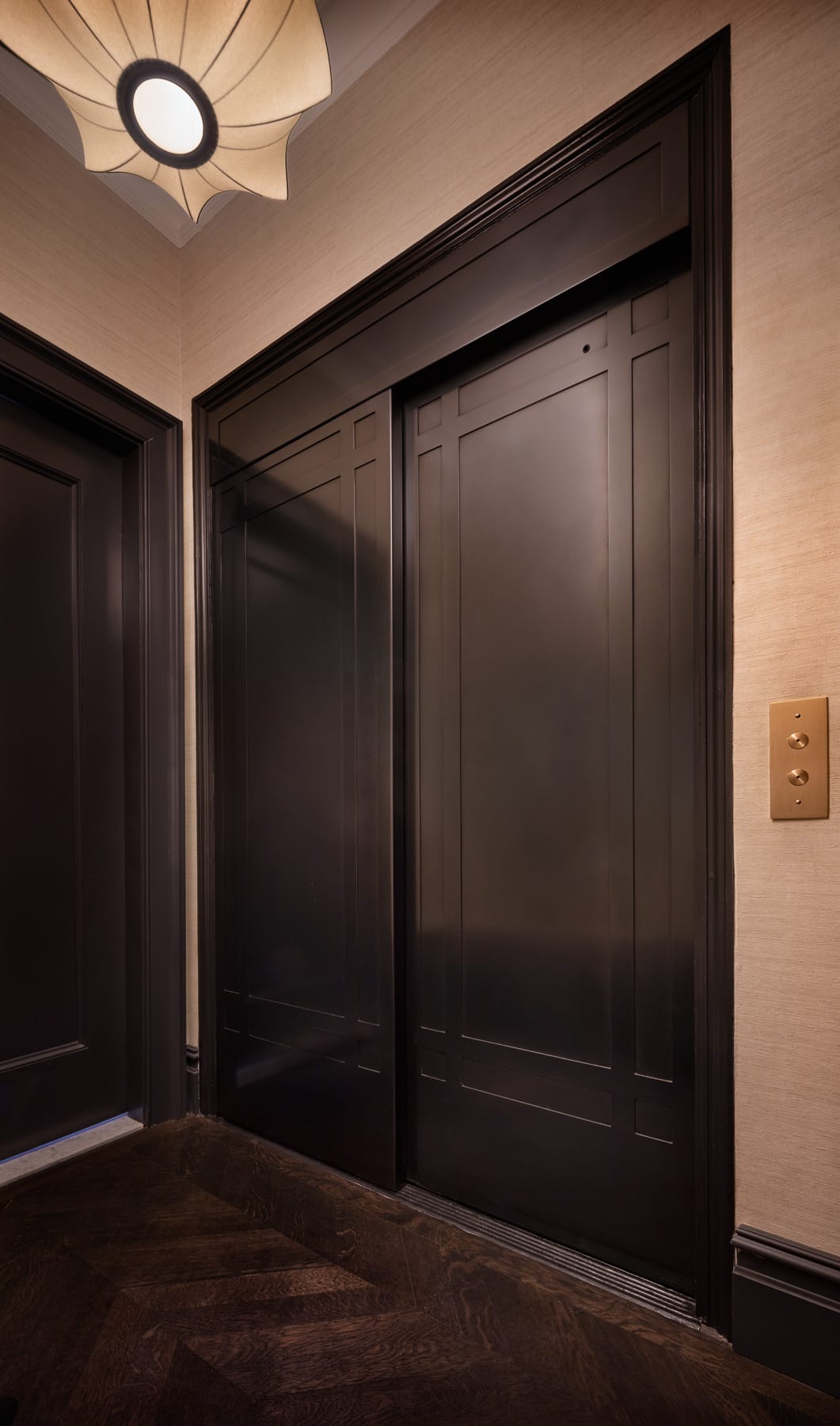 A closed dark-paneled elevator door is set in a hallway with beige walls, dark wood flooring, a ceiling light, and a brass call button.