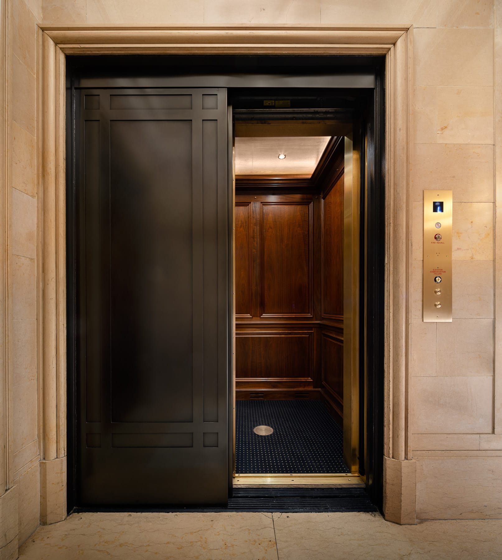 A partially open dark-paneled elevator reveals a wood-paneled interior, framed by light stone walls with a brass control panel.