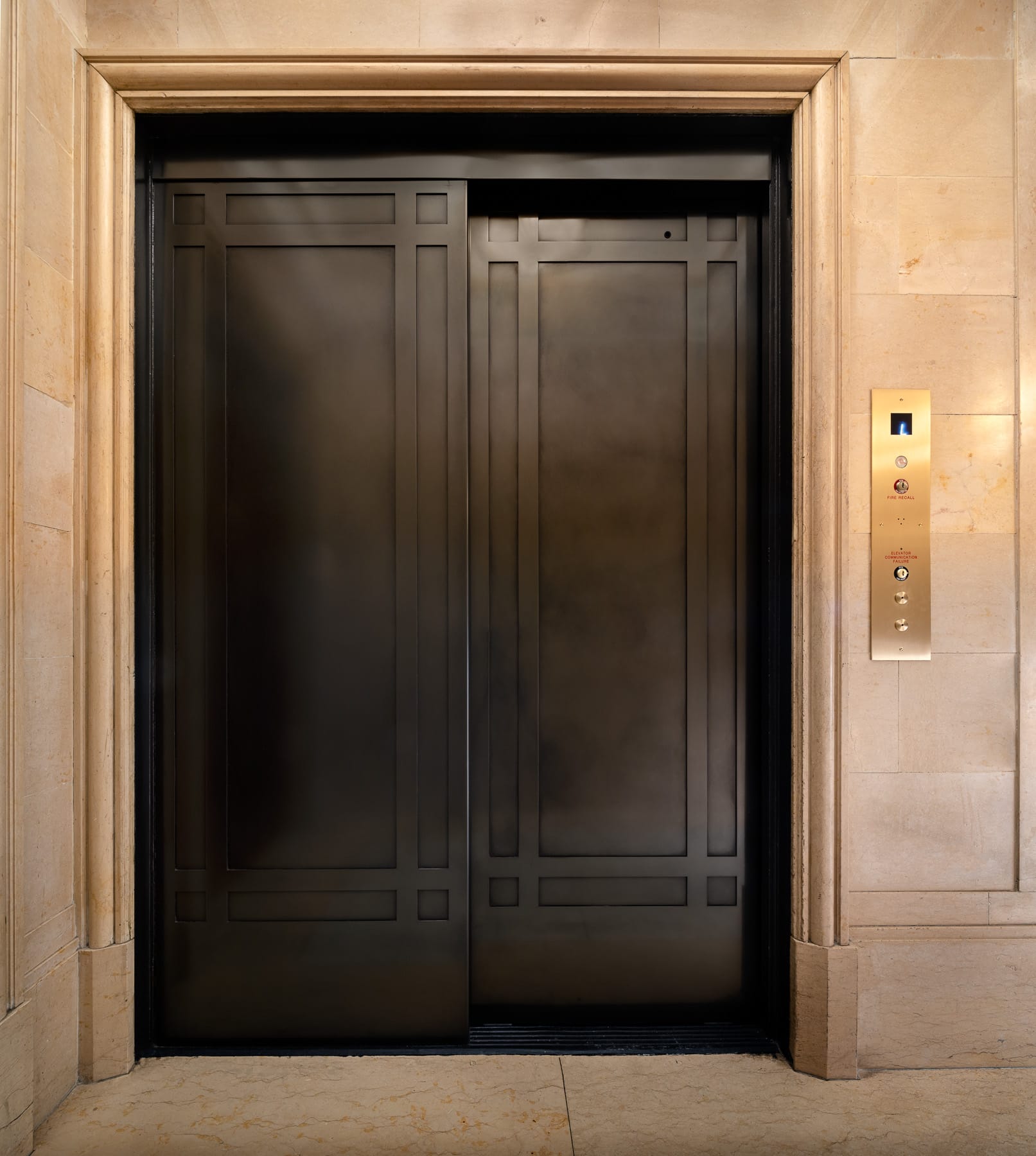 A closed dark-paneled elevator is framed by light stone walls beside a brass control panel displaying floor 1.