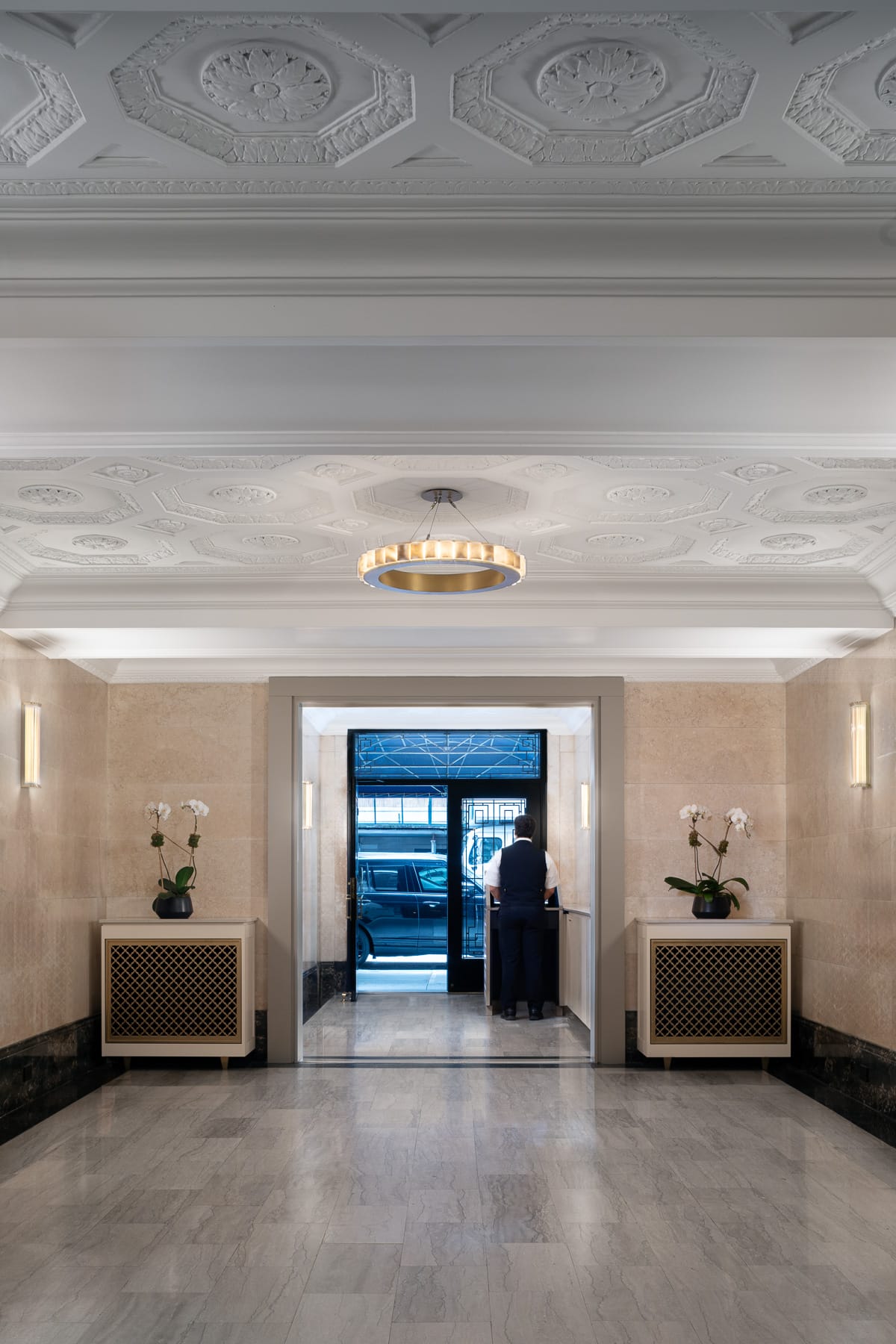 A marble-floored lobby with an ornate white ceiling, circular pendant light, orchid arrangements, and a uniformed attendant standing near the glass entrance.