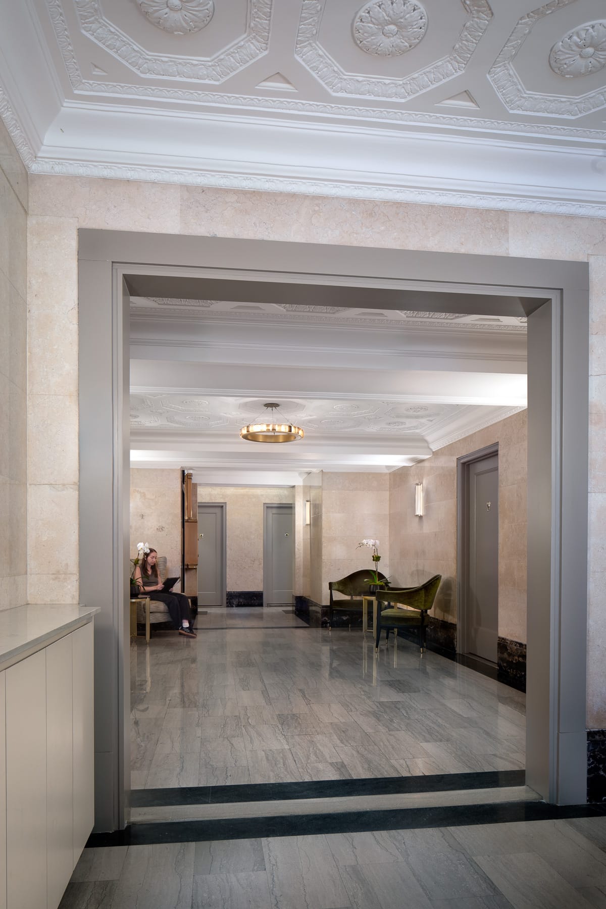 A marble-floored lobby viewed through a wide doorway has ornate ceilings, seating areas with orchids, and a person seated along the left wall.