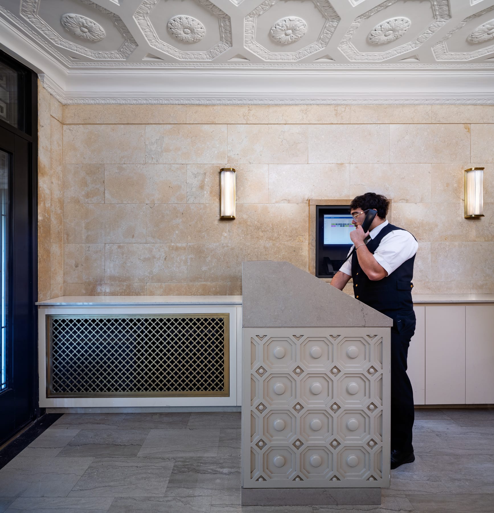 A uniformed attendant speaks on a telephone behind a decorative lobby desk beneath an ornate ceiling and wall sconces.