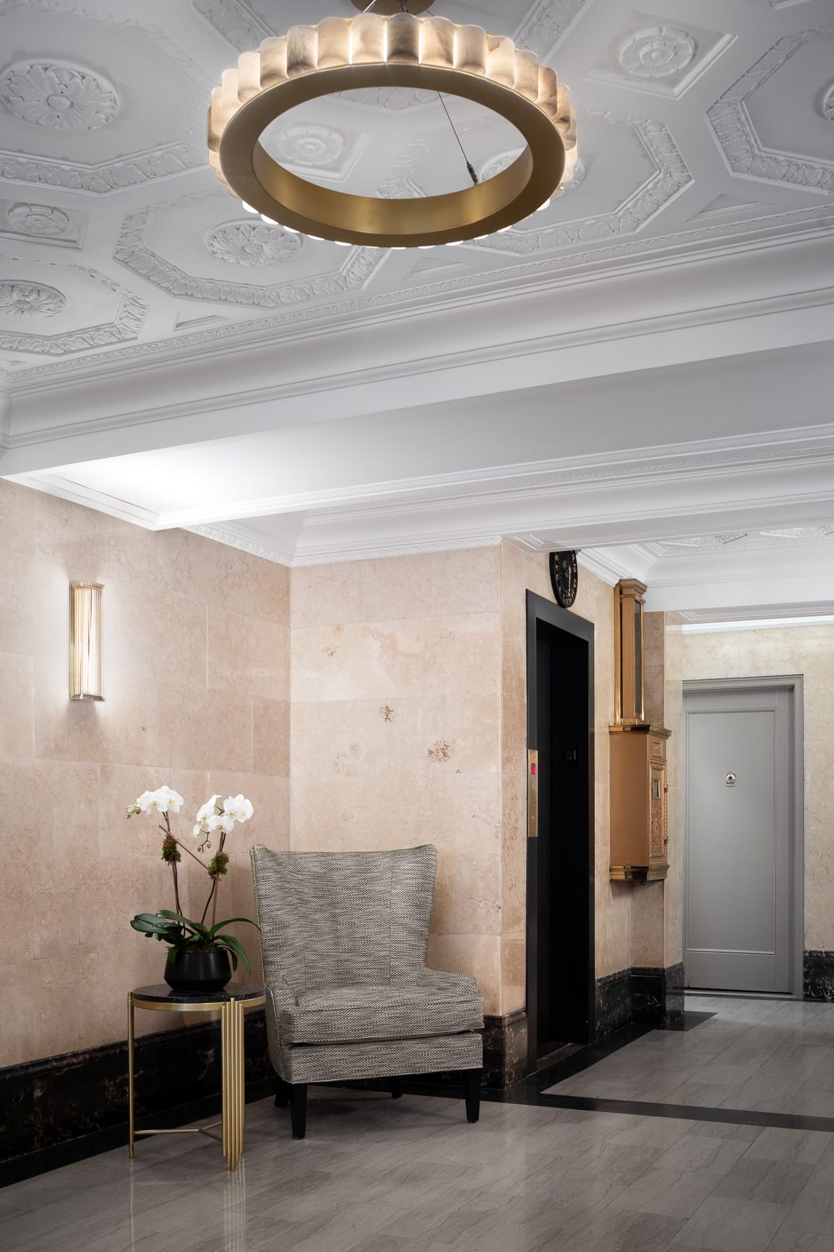A lobby seating area features a gray upholstered chair beside an orchid arrangement, with an elevator and gray door beneath an ornate white ceiling and circular pendant light.