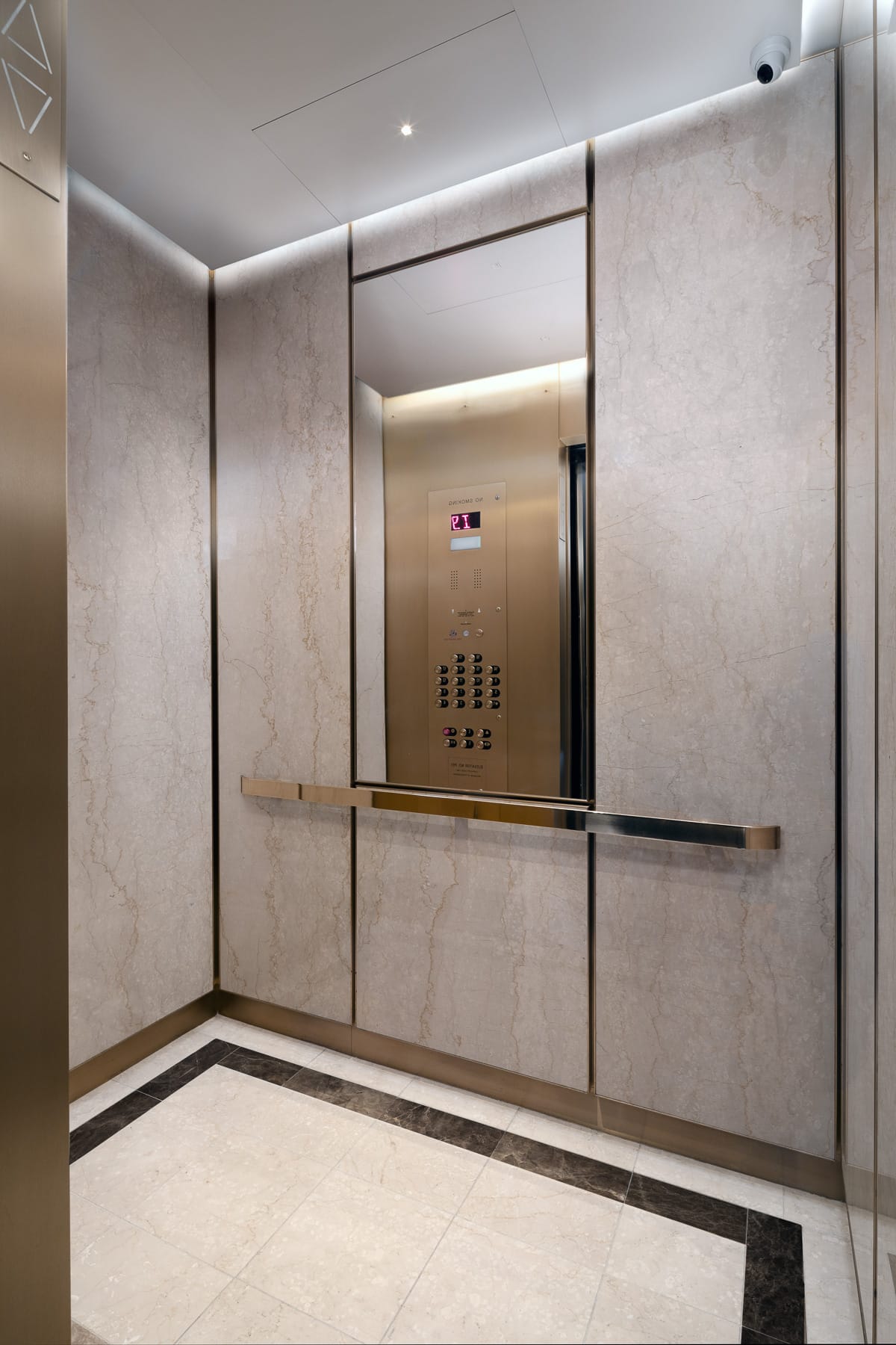 Elevator interior with light stone wall panels, a large mirror, brass-toned controls and handrail, tiled floor, recessed lighting, and a ceiling-mounted security camera.