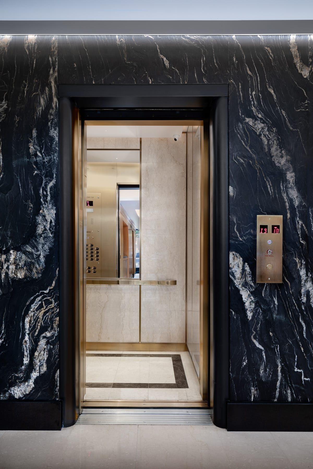 Open elevator doorway framed by black veined stone, revealing a light stone interior with brass-toned controls and handrail, with a floor indicator and call panel mounted beside the entrance.