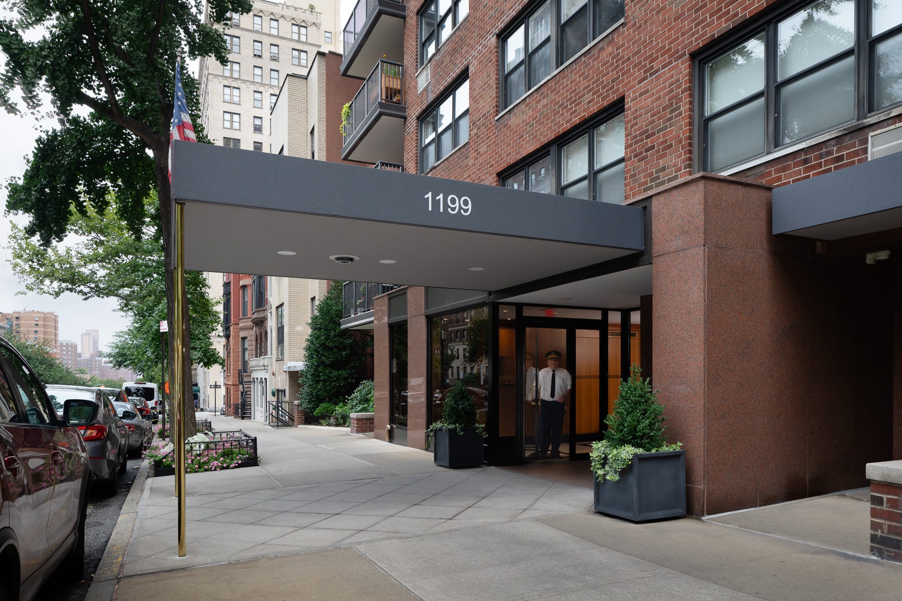 A red-brick residential building entrance with a canopy marked “1199,” a uniformed attendant by the glass doors, and parked cars along the tree-lined street.