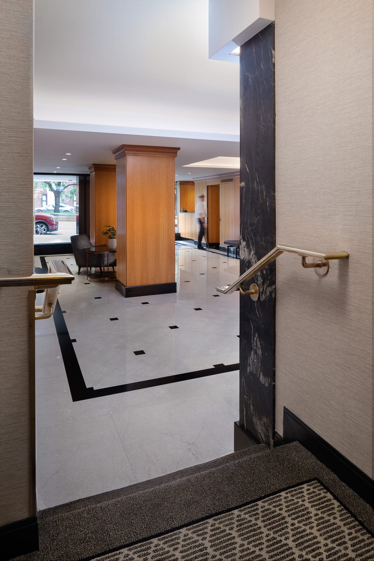 A short staircase with brass handrails overlooks a wood-paneled lobby where a person walks across the polished tile floor.