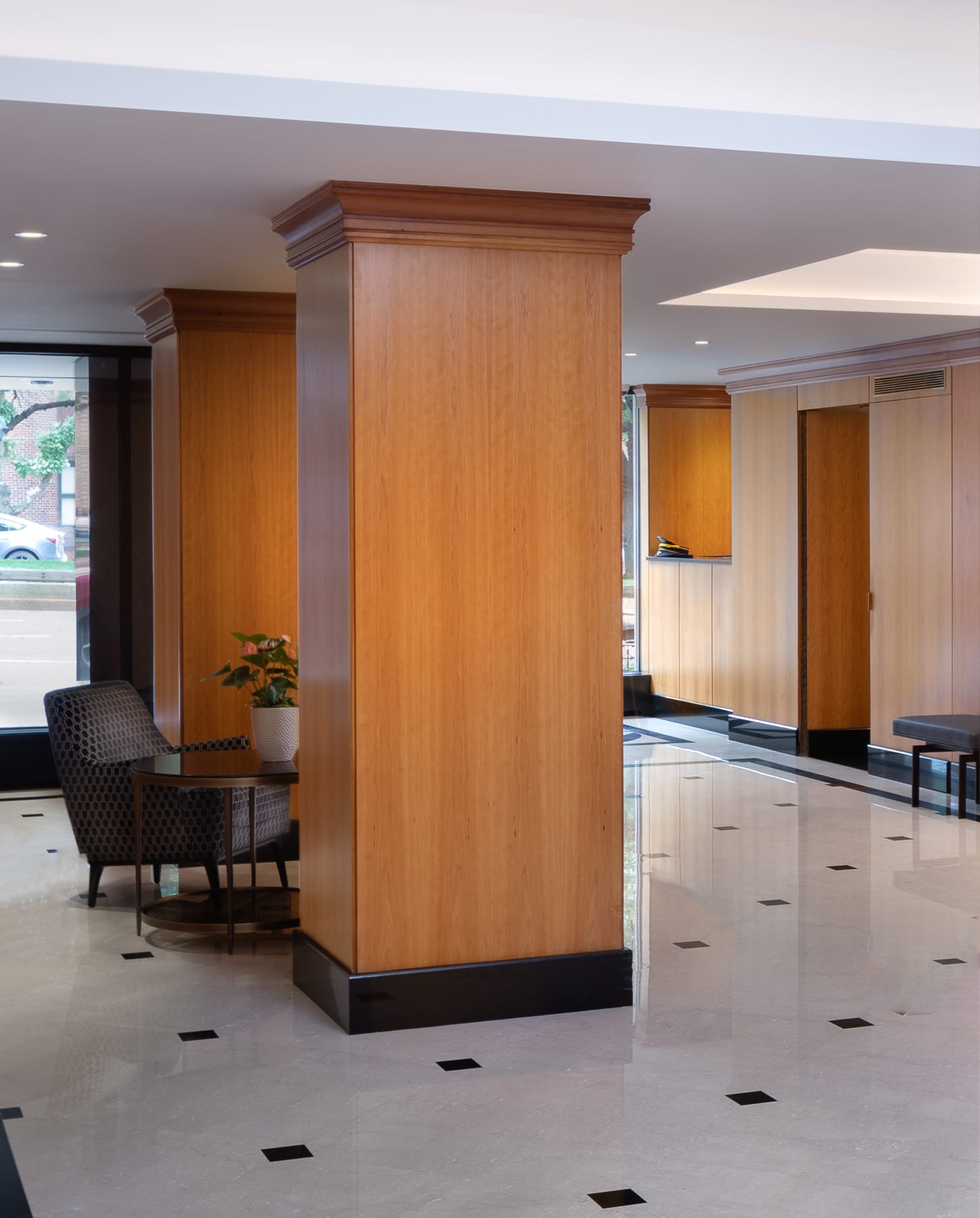 A residential lobby features large wood-paneled columns, polished tile flooring, a small seating area, and a reception counter near the windows.