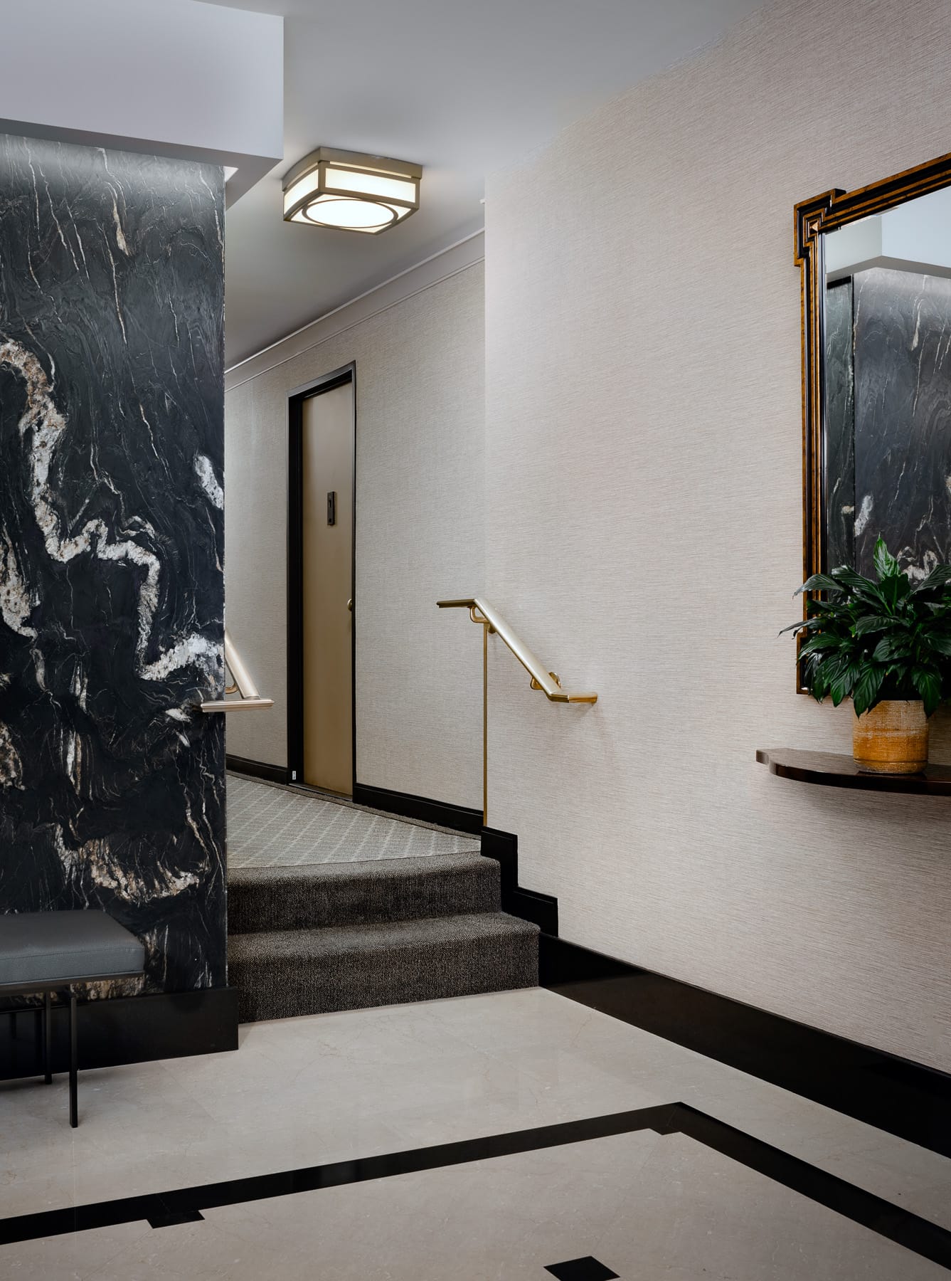 A carpeted stair and landing with brass handrails sits beside a black marble column, wall mirror, and potted plant.