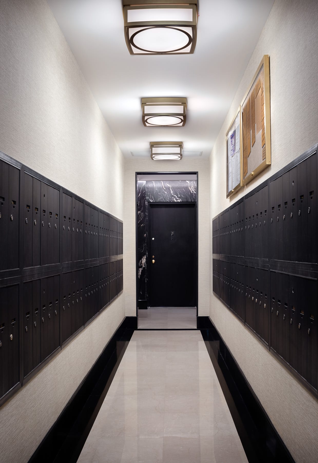 A narrow mailroom corridor has rows of dark mailboxes on both walls, overhead lights, and a black door at the far end.